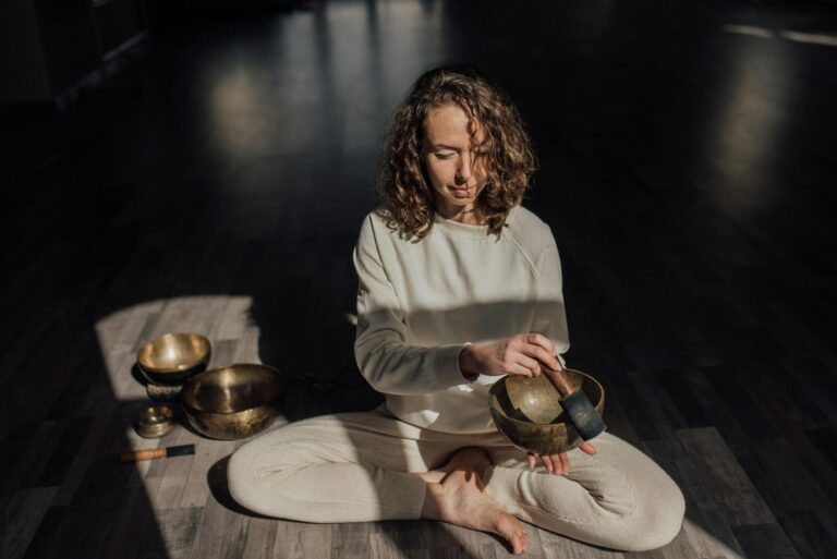 Female sound therapist sitting with crossed legs on parquet while playing singing bowl with mallet during meditation practice